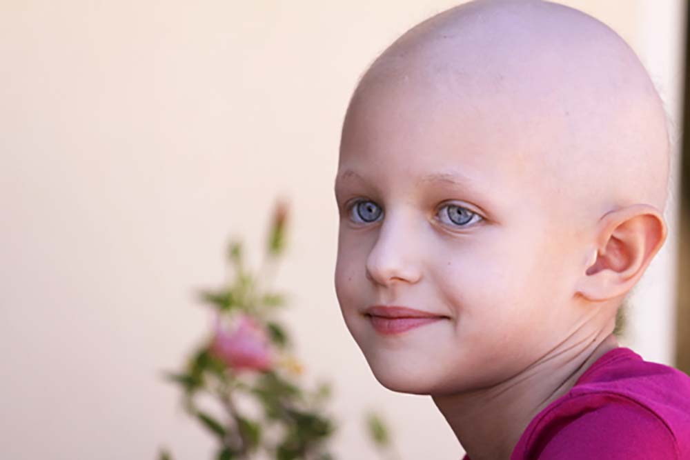 Een kind met een kaal hoofd van de chemotherapie met een roze shirt aan kijkt met een glimlach naar de linkerkant van het beeld. In de achtergrond is wazig de tak van een bloemenstruik te zien.