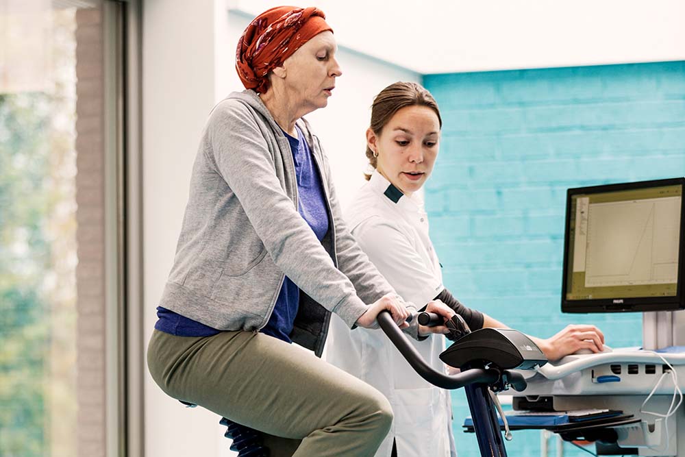 vrouw met kanker draagt hoofddoek en zit op een trainingsfiets. Een vrouwelijke zorgverlener staat naast haar en kijkt op het scherm van de fiets.