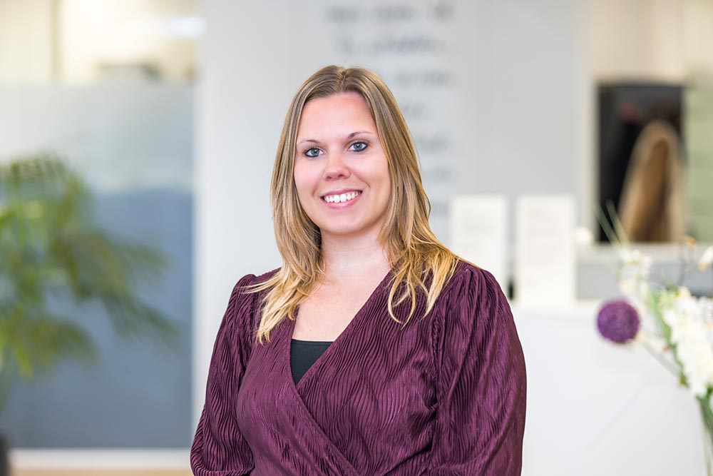 Eveline-IKNL portretfoto van Eveline, een witte vrouw met halflang blond haar. Ze draagt een paarse top en staat glimlachend voor een witte kantoorachtergrond.