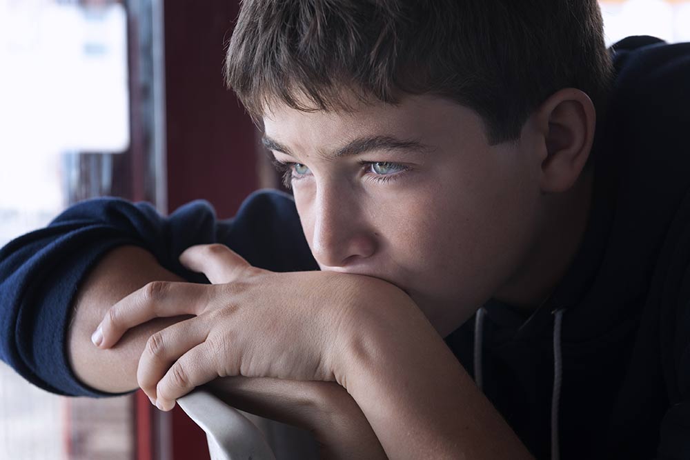 Een jongen van ongeveer 14 jaar met bruin haar, vanaf de schouders in beeld, staart peizend met het hoofd op de handen links naar links buiten beeld