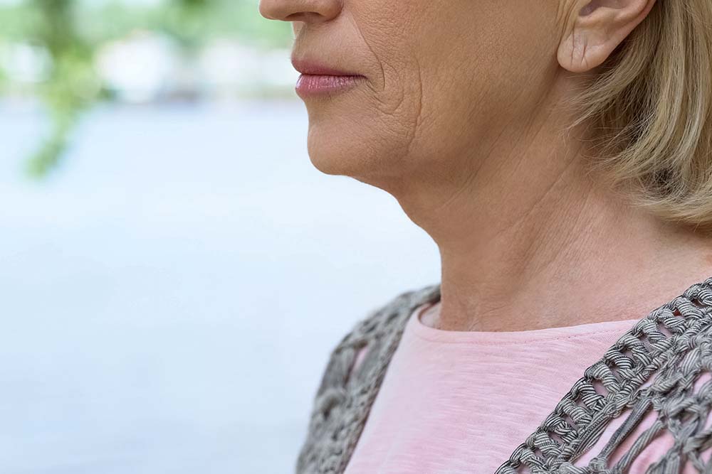 Zijaanzicht van blonde vrouw met roze shirt, van neus tot schouders in beeld. Het middelvoorste deel van de hals, waar de schildklier zit, is het focuspunt.