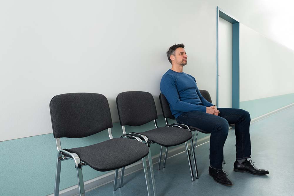 man met kort grijs haar en blauwe trui wacht op een stoel in een ziekenhuisgang. Naast hem staan nog 2 lege stoelen.