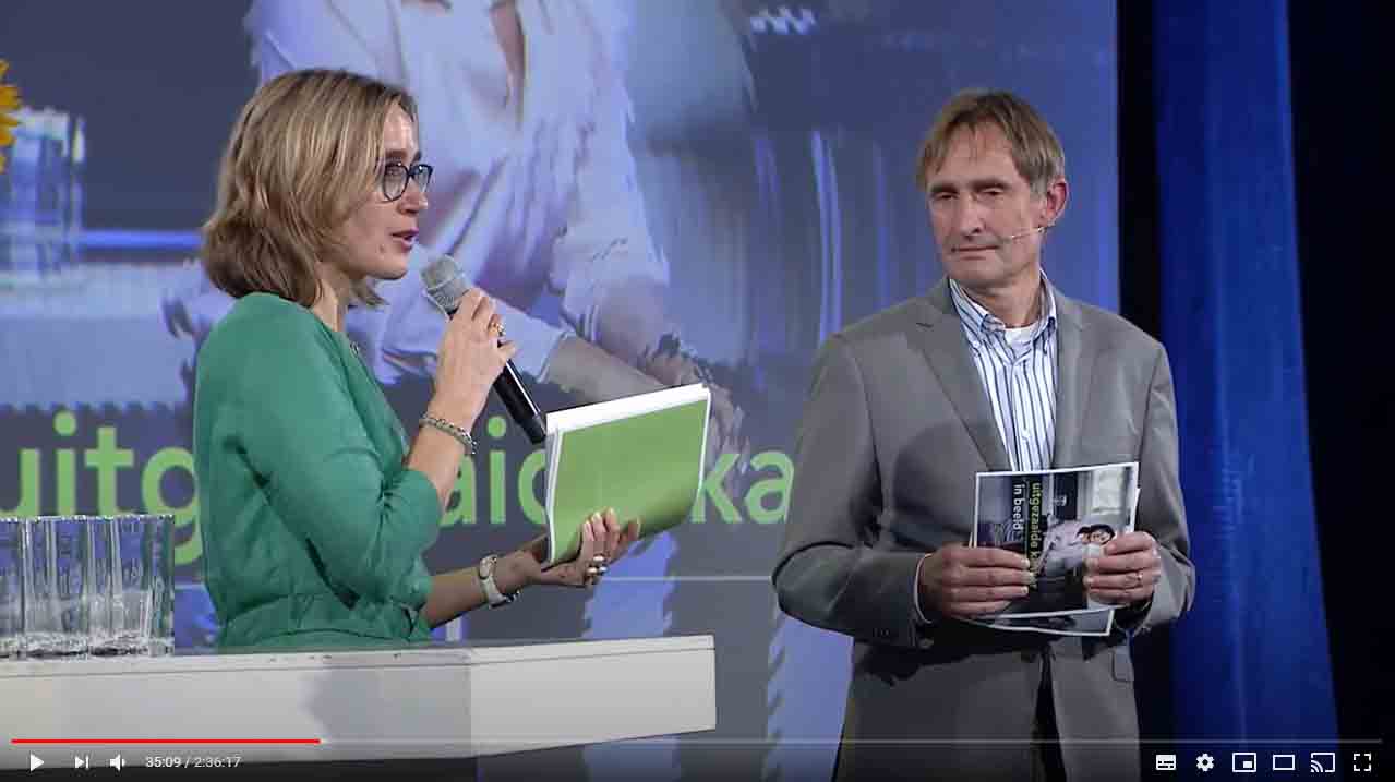 Dorien Tange, een blonde vrouw met een bril en een groene jurk, spreekt in een microfoon en heeft het rapport 'Uitgezaaide kanker in beeld' vast. Thijs Merkx, een grijze man, staat rechts van haar.