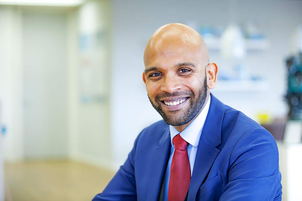 Portretfoto van Avinash, een bruine man met kaal hoofd en korte zwarte baard. Hij draagt een blauw pak met witte stropdas en kijkt glimlachend de camera in.