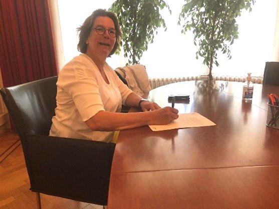 voorzitter Carla van Herpen, een witte vrouw met donker kort haar en een bril, zit aan tafel en ondertekend een document
