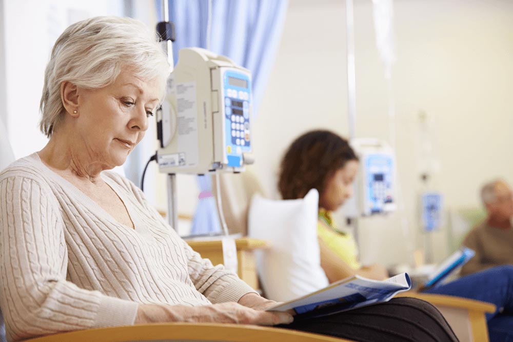 twee vrouwen in dagbehandelingsetting, zitten op stoelen en lezen een boekje. vrouw in de voorgrond heeft kort wit haar, vrouw in de achtergrond heeft bruin kroeshaar.