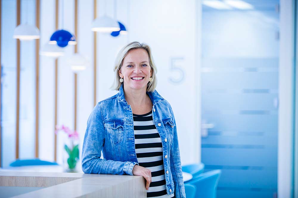 Lonneke, een witte vrouw met een korte blonde bob, staat glimlachend met haar arm geleund op een bureau. Ze draagt een spijkerjasje en een zwart-wit gestreepte top