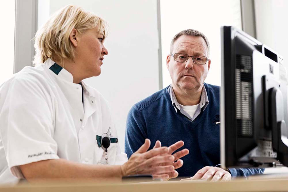 Een vrouwelijke arts met kort blond haar is in gesprek met man met bril, grijs kort haar en baard. Ze zitten links in beeld en de man kijkt op een beeldscherm dat rechts van achteren te zien is.